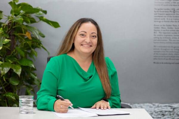 A woman in a green blouse sits at a desk signing papers, reflecting on Queensland's proposed reforms to enhance tenants' rights. A plant and a glass of water beside her symbolize the fresh growth and safety standards these changes aim to nurture.
