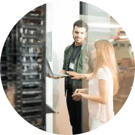 Two people stand near a server rack. One holds a laptop, and they appear to be discussing something technical.